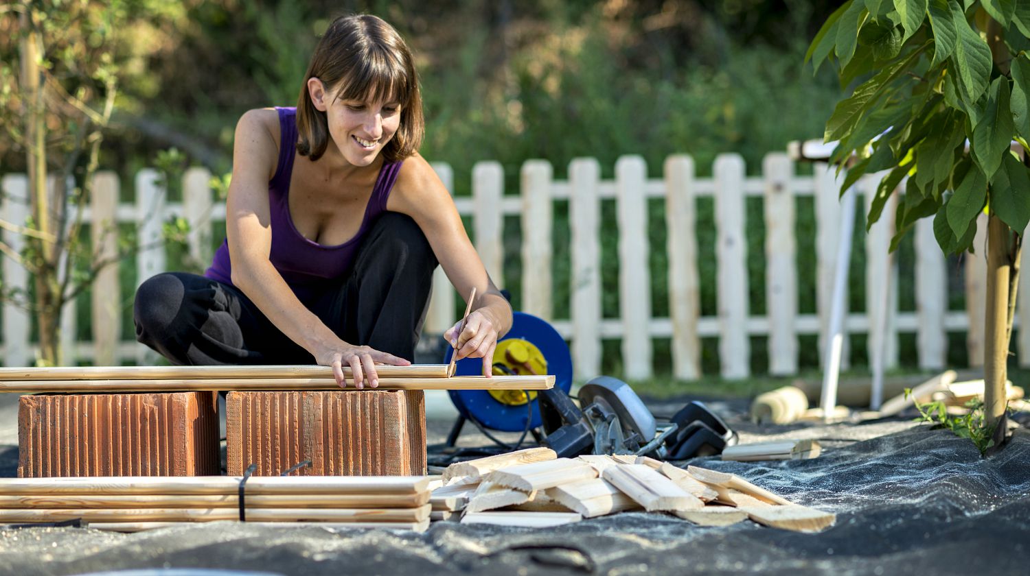 Smiling young woman marking with a pencil where to cut a wooden plank for a backyard fence | Backyard Projects To Enjoy Time Outdoors This Summer | Featured