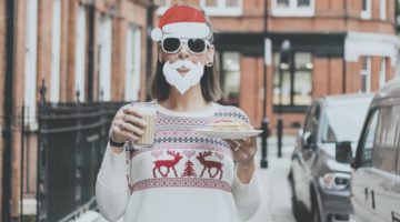 woman wearing an ugly Christmas sweater with mask santa hat and beard holding drink and slice of cake in the street | Christmas Sweater Ideas You Can DIY On A Budget | make your own christmas sweater online | Featured