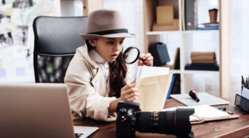 Little girl in cloak and hat is sitting at desk looking at photos with magnifying glass | How To Make Spy Gear (For Adults And Kids) | spy stuff | Featured