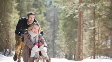 man-pushing-woman-on-sled-in-snowy-woods | How To Make A Wooden Sled Your Kids Can Enjoy This Winter | Featured