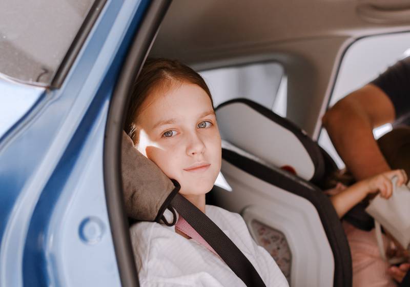 Little girl sitting in opened car and fastening seat belt with soft grey pillow | Seat Belt Pillows