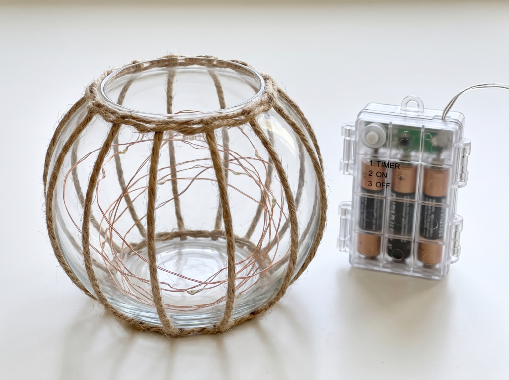 Clear battery box with three AA batteries visible sitting behind an unlit twine-wrapped glass bowl with fairy lights coiled inside