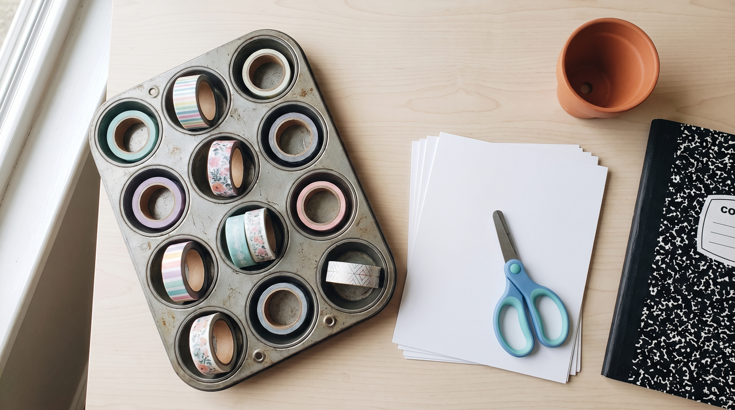 Washi tape rolls sorted in a muffin tin with cardstock, kid-safe scissors and a notebook on a craft table