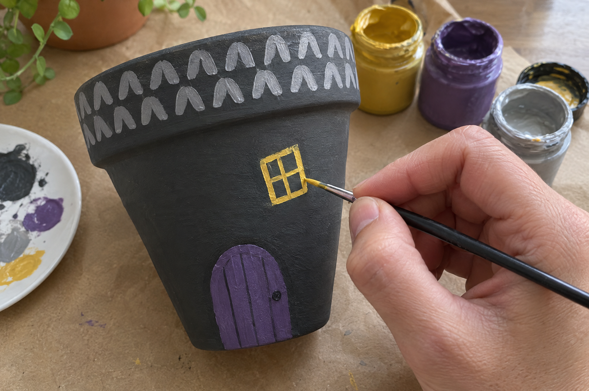 Painting a yellow window and a shingle roofline on a dark gray terracotta pot haunted house